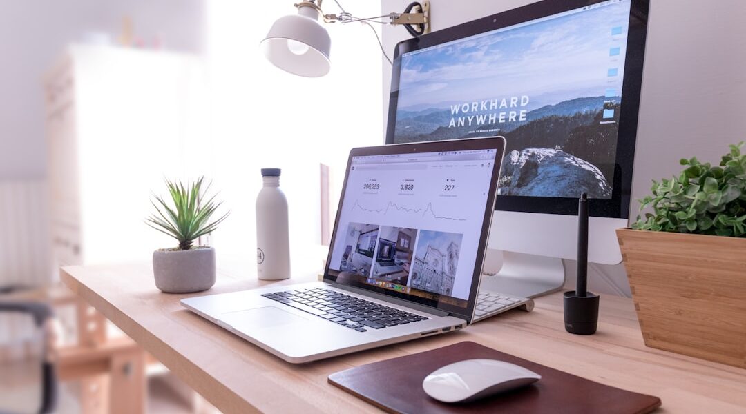A workspace dedicated to digital marketing features a laptop, desktop monitor, white desk lamp, potted plant, water bottle, pen tablet, and a wireless mouse on a wooden desk. The monitor displays text "WORKHARD ANYWHERE".