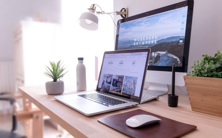 A workspace dedicated to digital marketing features a laptop, desktop monitor, white desk lamp, potted plant, water bottle, pen tablet, and a wireless mouse on a wooden desk. The monitor displays text "WORKHARD ANYWHERE".