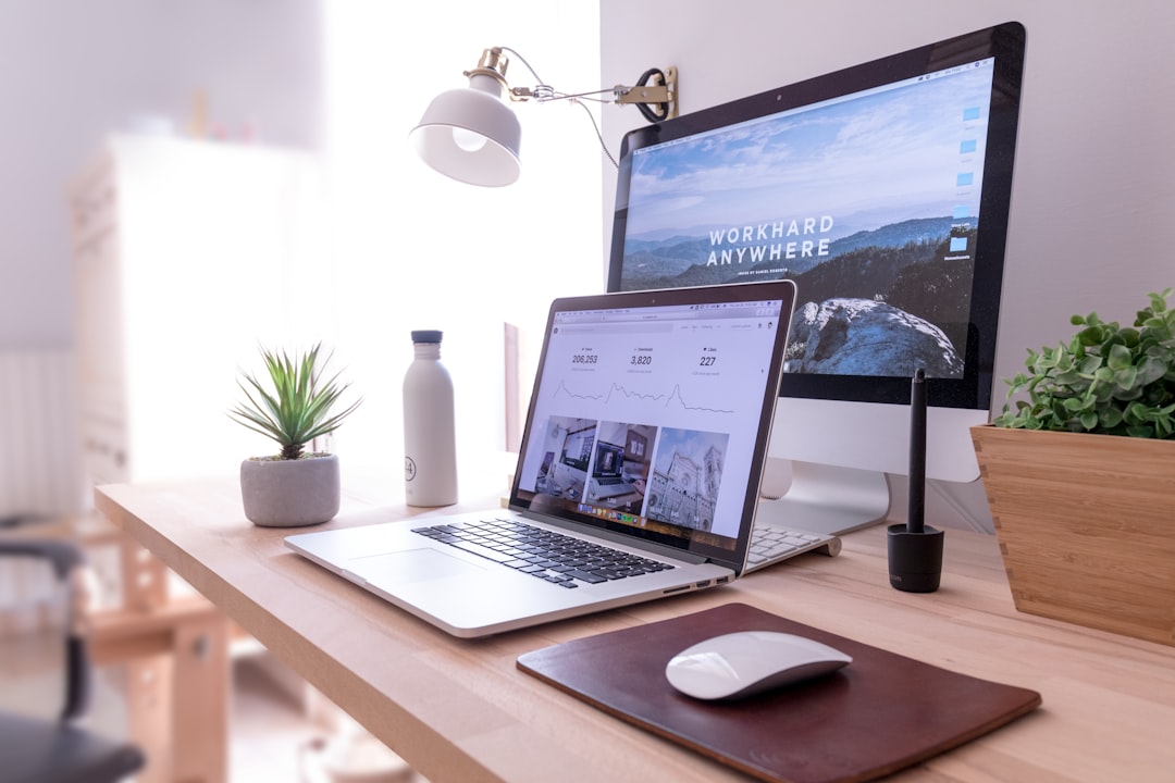 A workspace dedicated to digital marketing features a laptop, desktop monitor, white desk lamp, potted plant, water bottle, pen tablet, and a wireless mouse on a wooden desk. The monitor displays text "WORKHARD ANYWHERE".