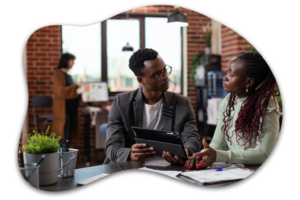 Two people are sitting and discussing something, with one holding a tablet. Another person works at a desk in the background. The setting is an office with brick walls and modern decor.