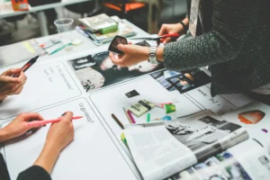 Three people work together at a table covered with magazines, markers, paper, and craft supplies, possibly creating a collage or vision board.