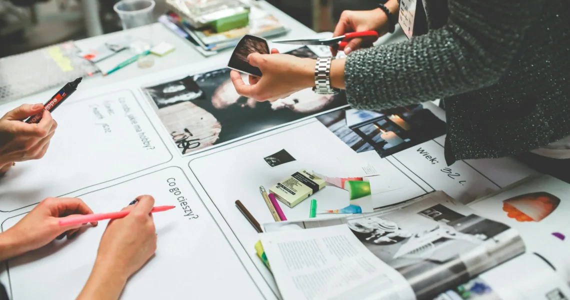 Three people work together at a table covered with magazines, markers, paper, and craft supplies, possibly creating a collage or vision board.