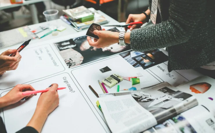 Three people work together at a table covered with magazines, markers, paper, and craft supplies, possibly creating a collage or vision board.