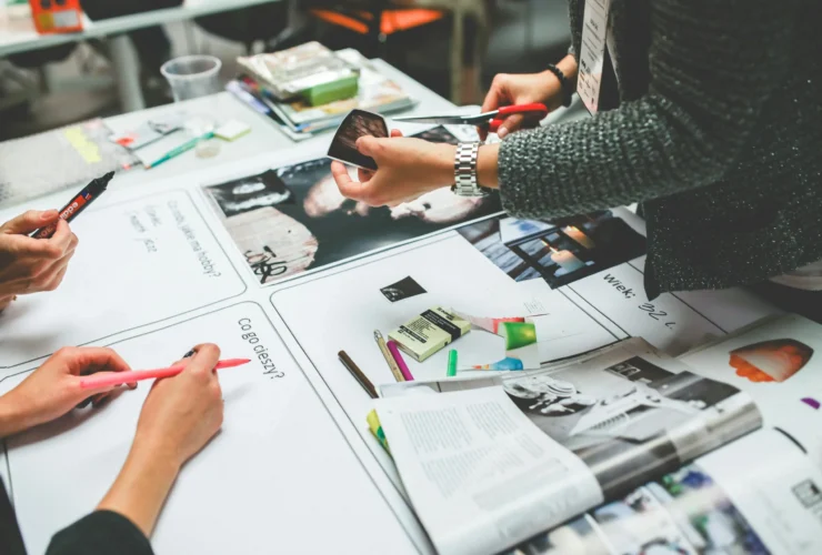 Three people work together at a table covered with magazines, markers, paper, and craft supplies, possibly creating a collage or vision board.