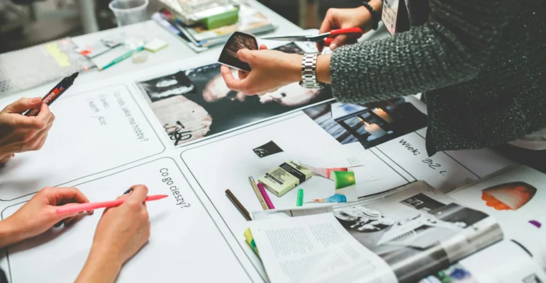 Three people work together at a table covered with magazines, markers, paper, and craft supplies, possibly creating a collage or vision board.
