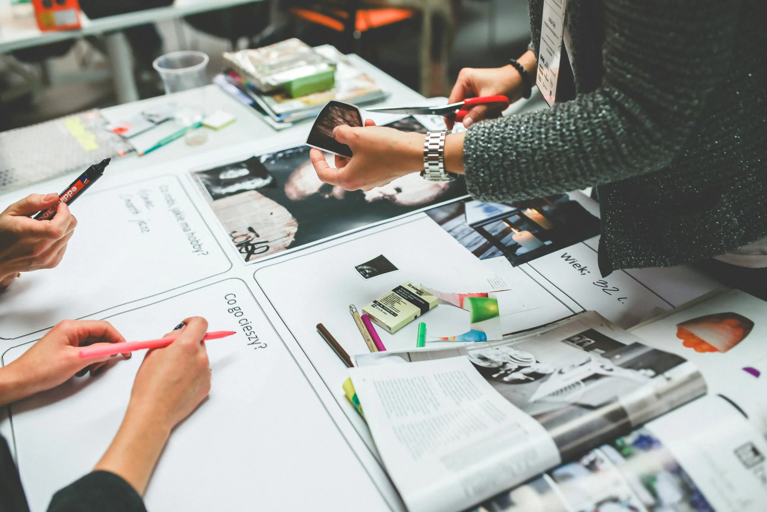Three people work together at a table covered with magazines, markers, paper, and craft supplies, possibly creating a collage or vision board.