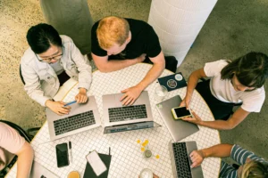 Four people sit around a round table working on laptops and a smartphone, viewed from above. Notebooks, pens, and drinks are also on the table.