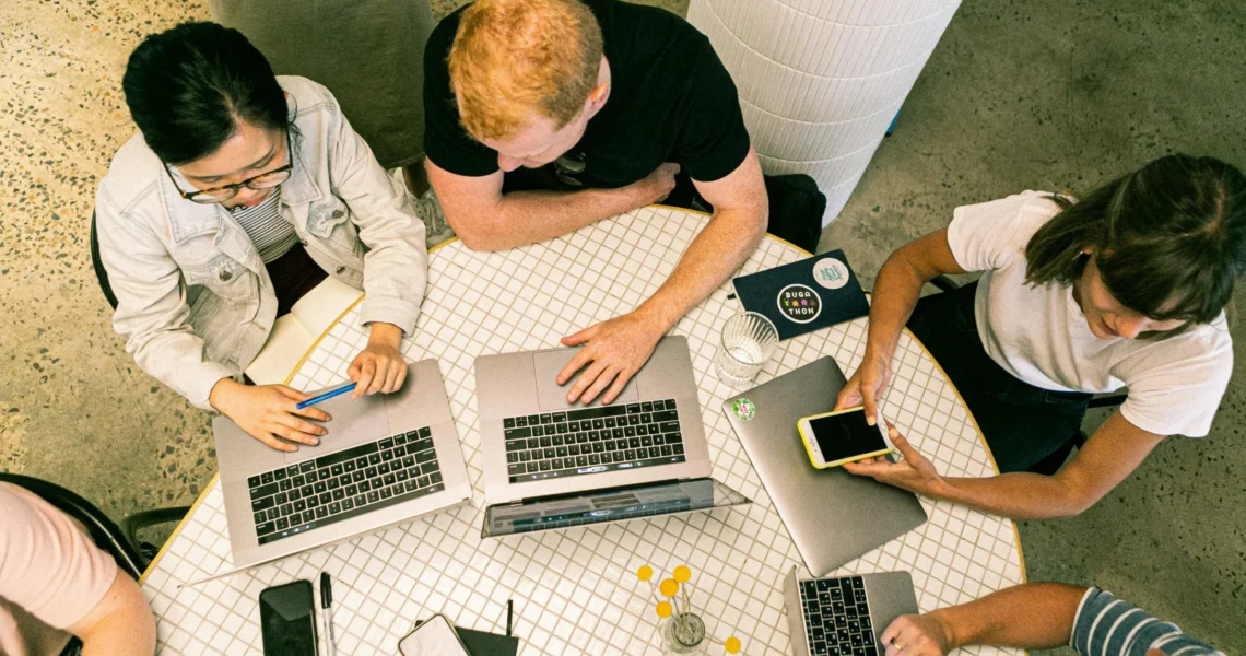 Four people sit around a round table working on laptops and a smartphone, viewed from above. Notebooks, pens, and drinks are also on the table.
