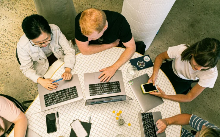 Four people sit around a round table working on laptops and a smartphone, viewed from above. Notebooks, pens, and drinks are also on the table.