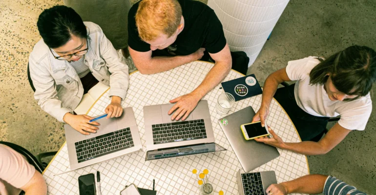 Four people sit around a round table working on laptops and a smartphone, viewed from above. Notebooks, pens, and drinks are also on the table.
