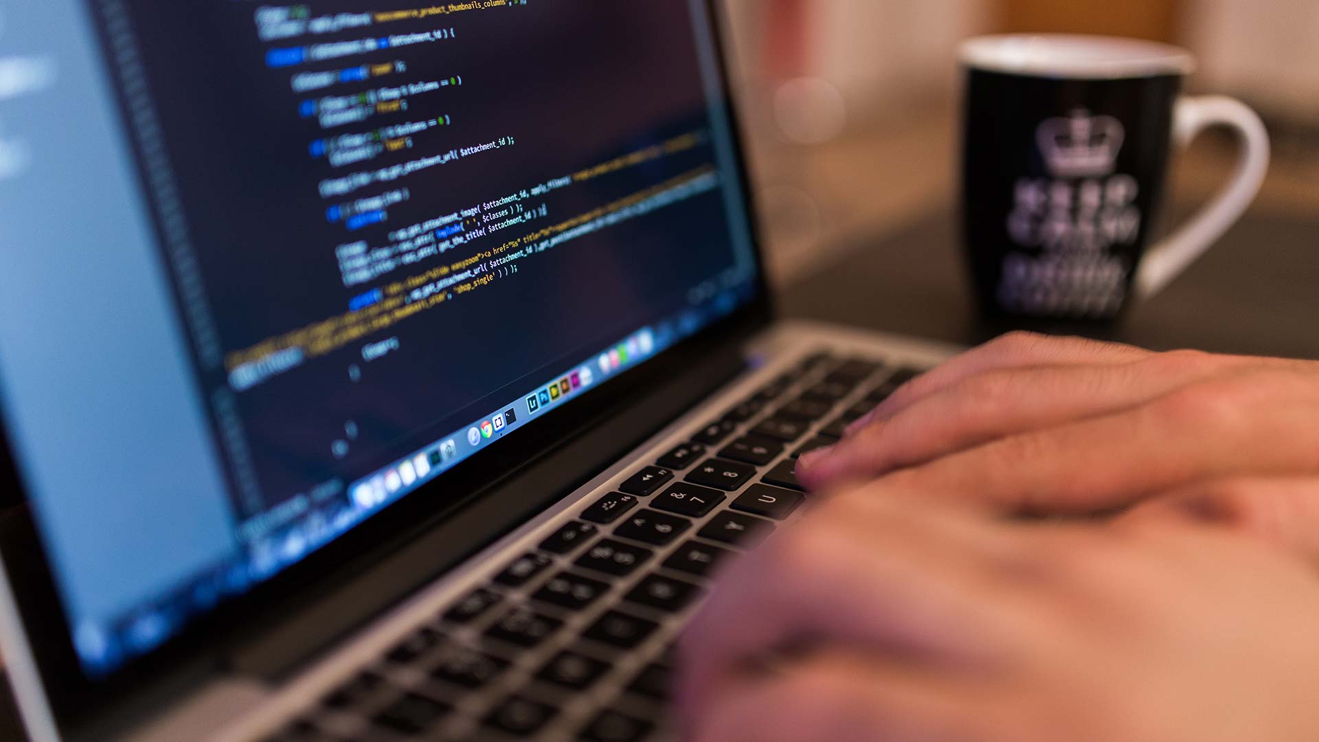 A person types on a laptop keyboard with computer code displayed on the screen; a coffee mug is visible in the background.