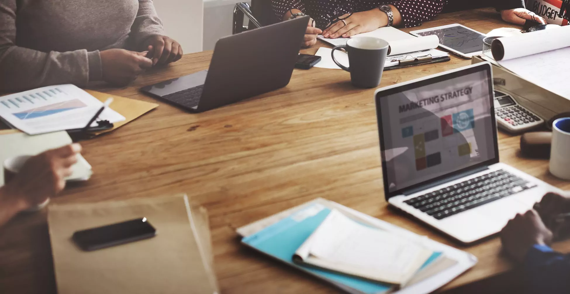 Four people sit at a wooden table with laptops, documents, coffee mugs, and a calculator, engaged in a work meeting. A laptop screen shows a marketing strategy presentation.
