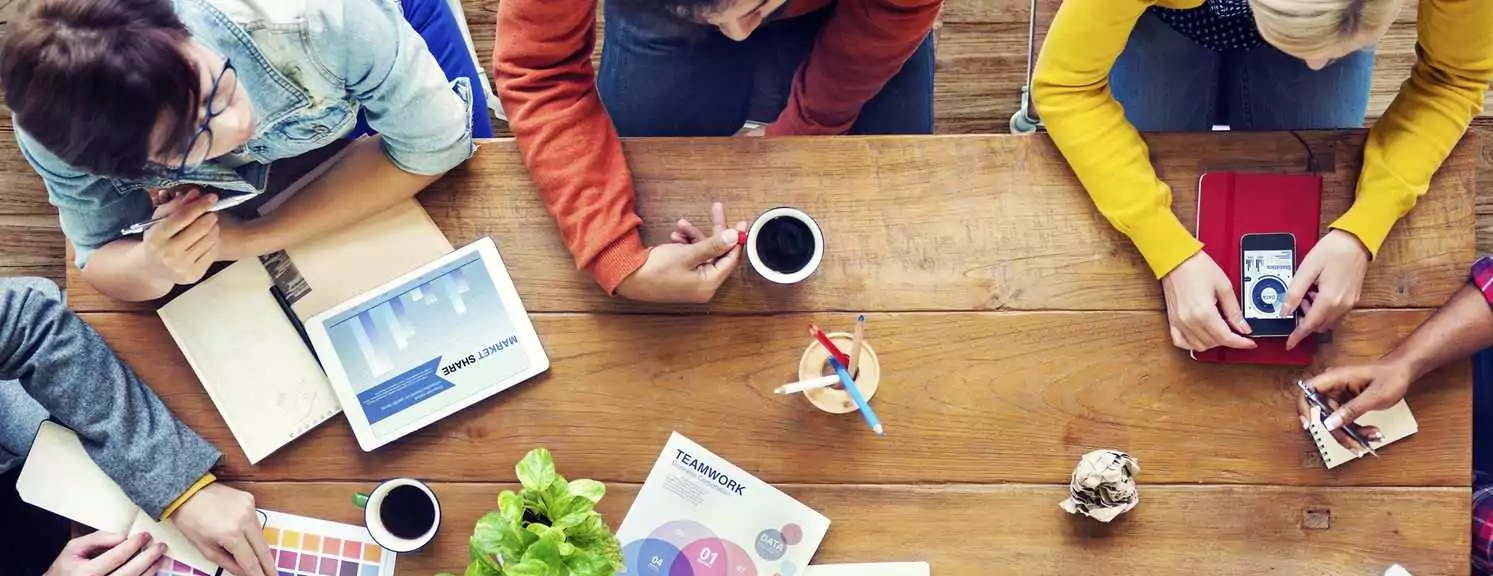 Four people sit around a wooden table with notebooks, a tablet, coffee cups, and documents, engaged in a collaborative meeting or discussion.