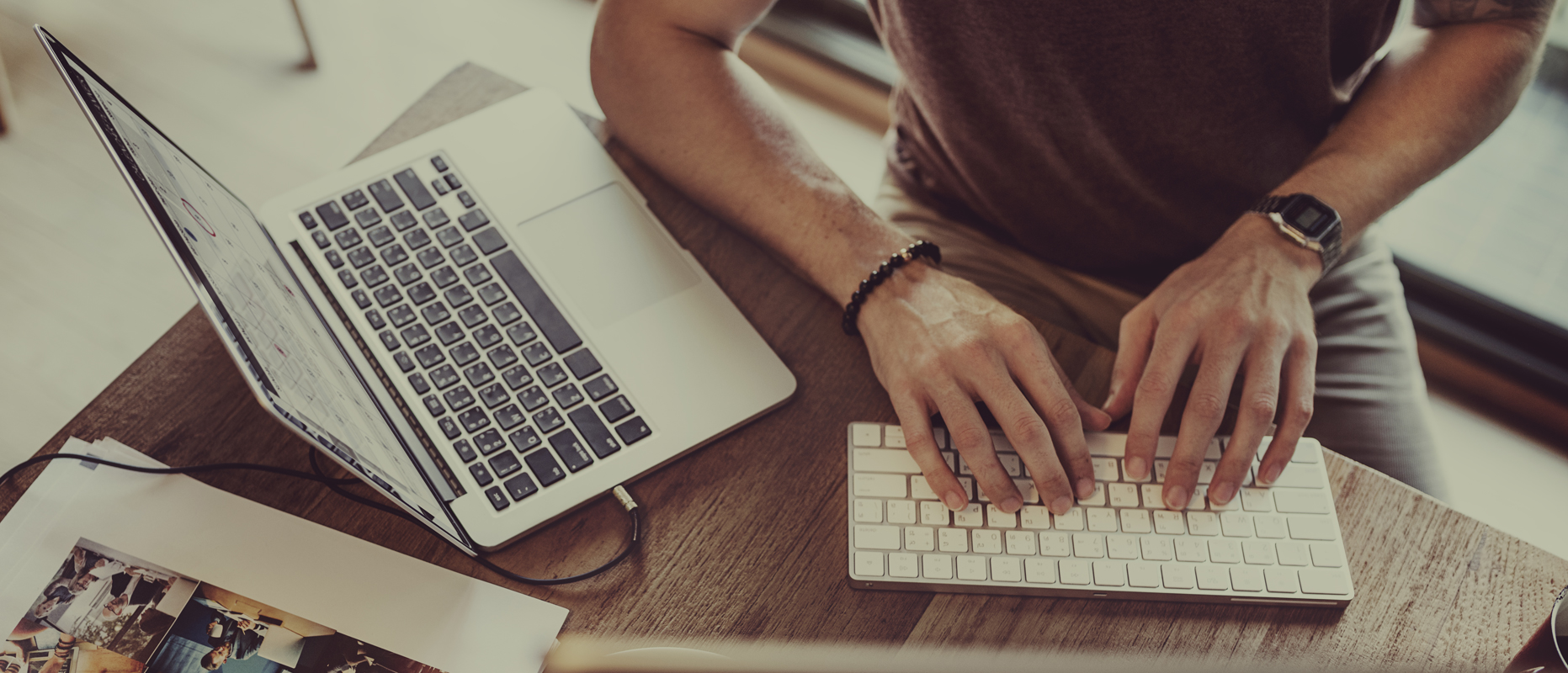 Person typing on a wireless keyboard next to a laptop on a wooden desk with papers and photos scattered nearby.