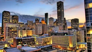 City skyline at sunset with illuminated office buildings, tall skyscrapers, and a partly cloudy sky in the background.