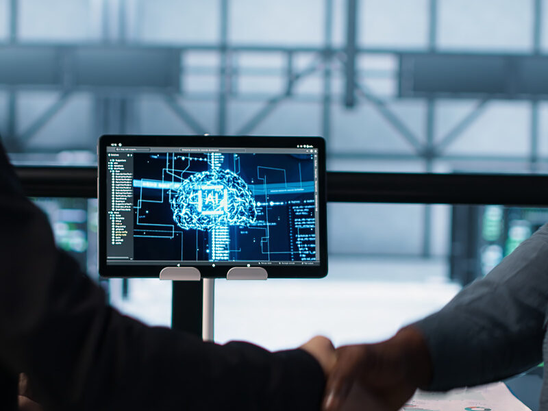 Two people shaking hands in front of a computer screen displaying an artificial intelligence graphic and data charts in a modern office setting, highlighting how artificial intelligence affects business operations and decision-making.