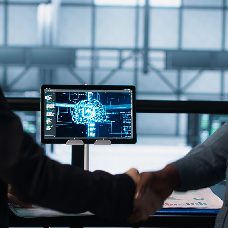 Two people shaking hands in front of a computer screen displaying an artificial intelligence graphic and data charts in a modern office setting, highlighting how artificial intelligence affects business operations and decision-making.