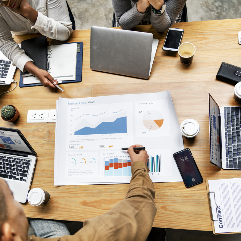 Three people collaborate around a wooden table with laptops, smartphones, and a large business chart showing graphs and data.